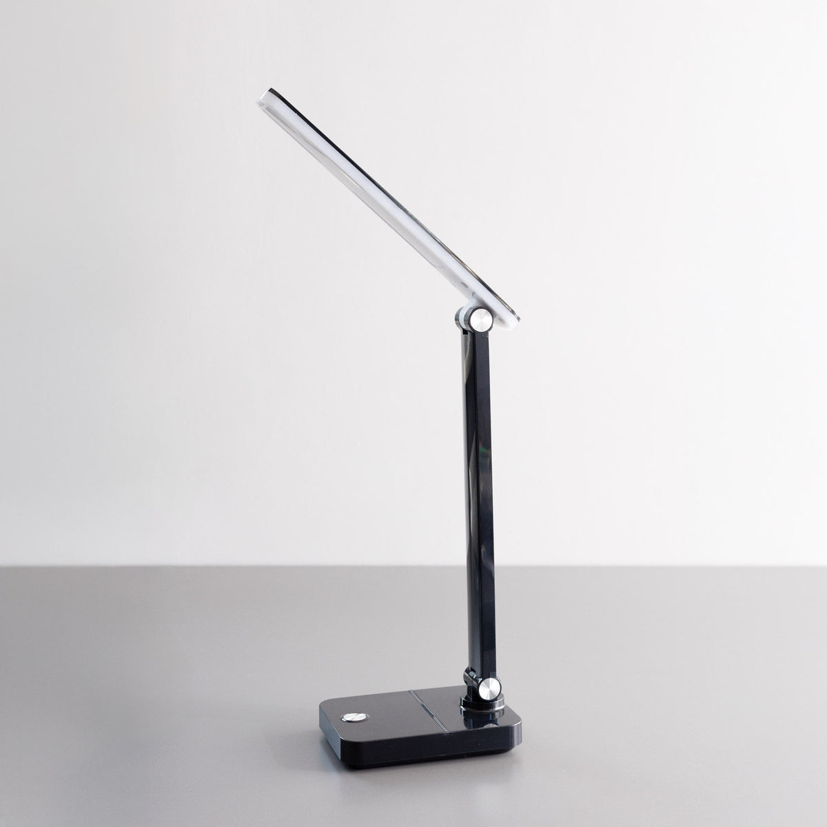 Même lampe de bureau noire éteinte, tête ajustable, base plate sur surface grise.