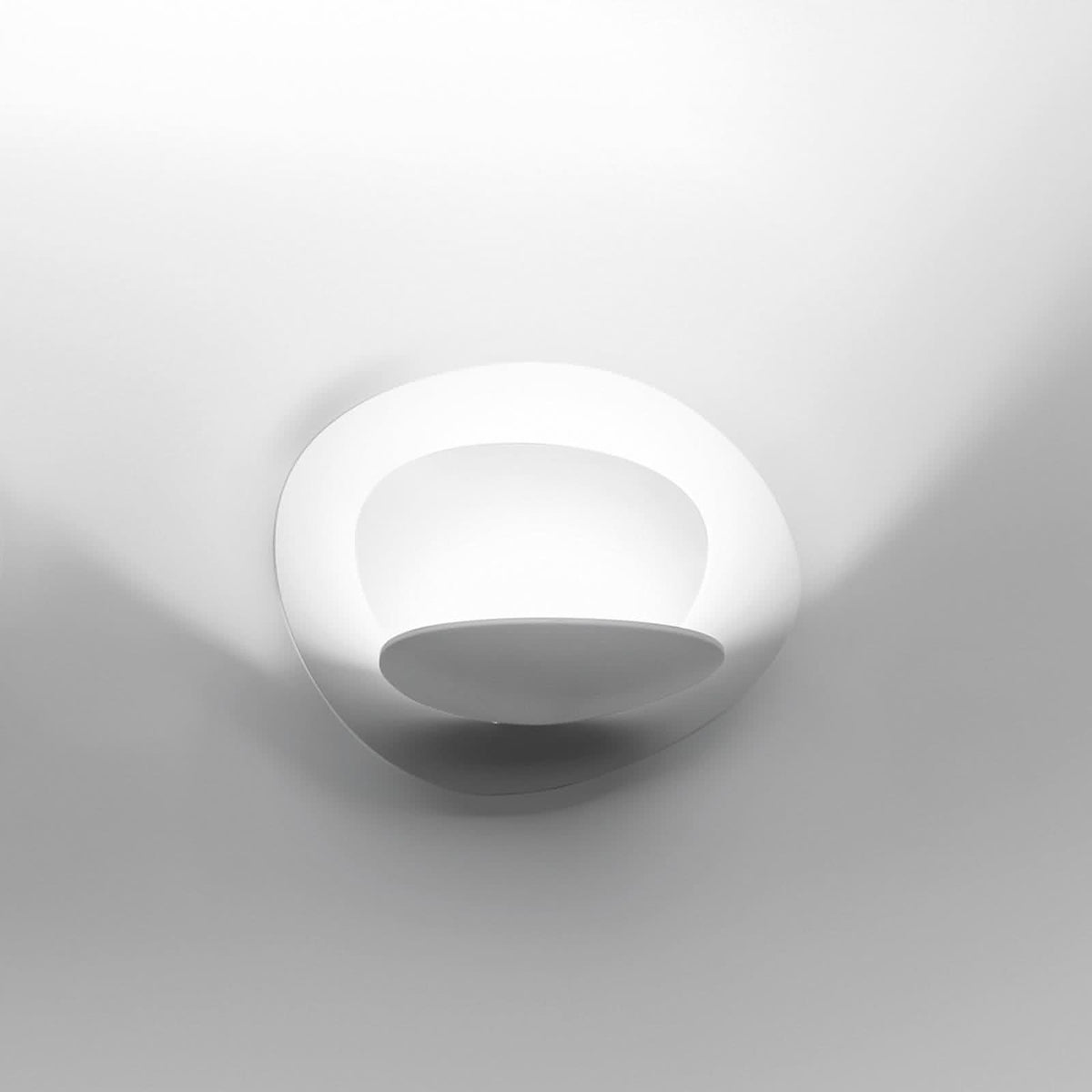 Applique murale blanche allumée, anneau ovalisé et halo lumineux sur le mur.