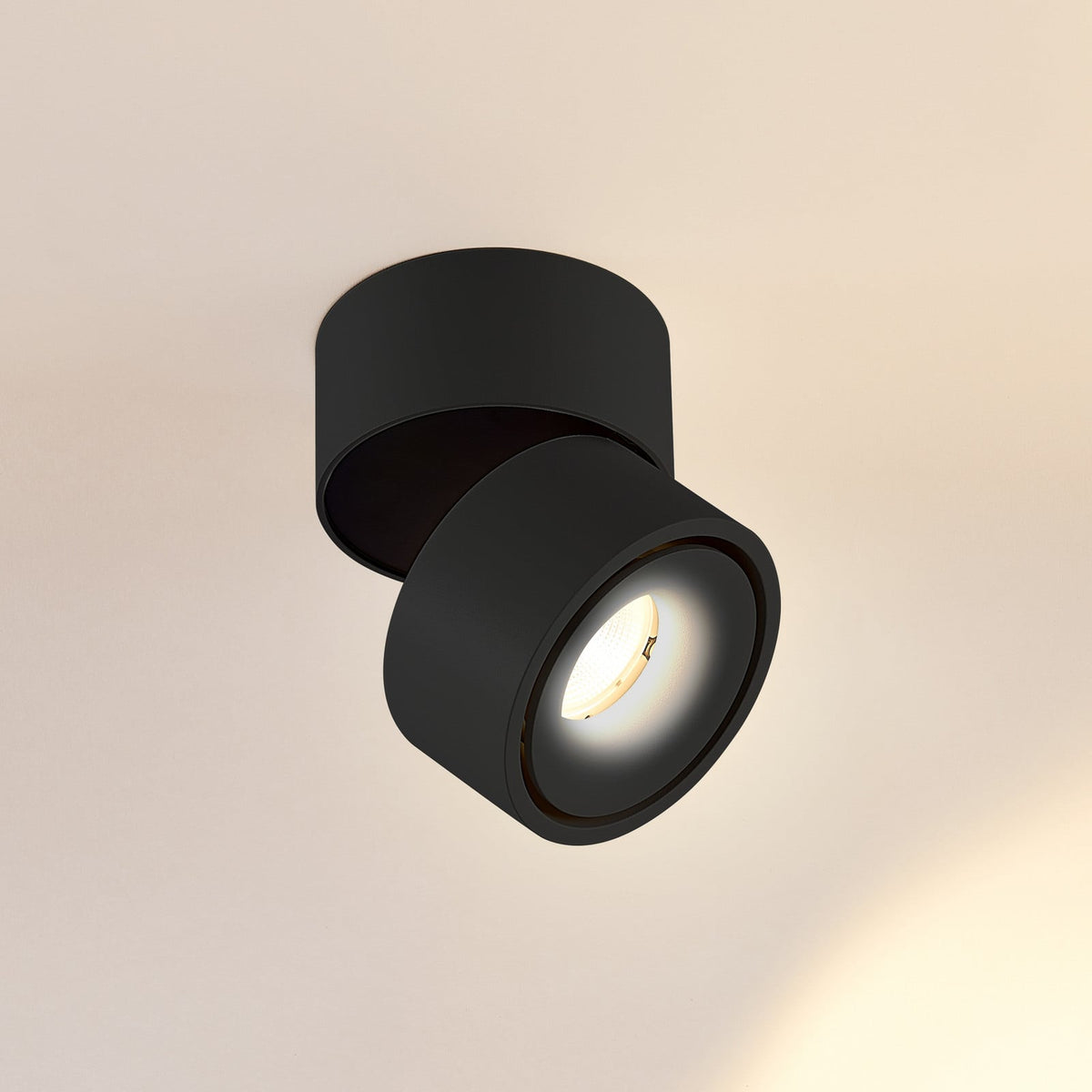 Spot LED noir orientable vu de trois quarts, allumé sur fond beige clair.