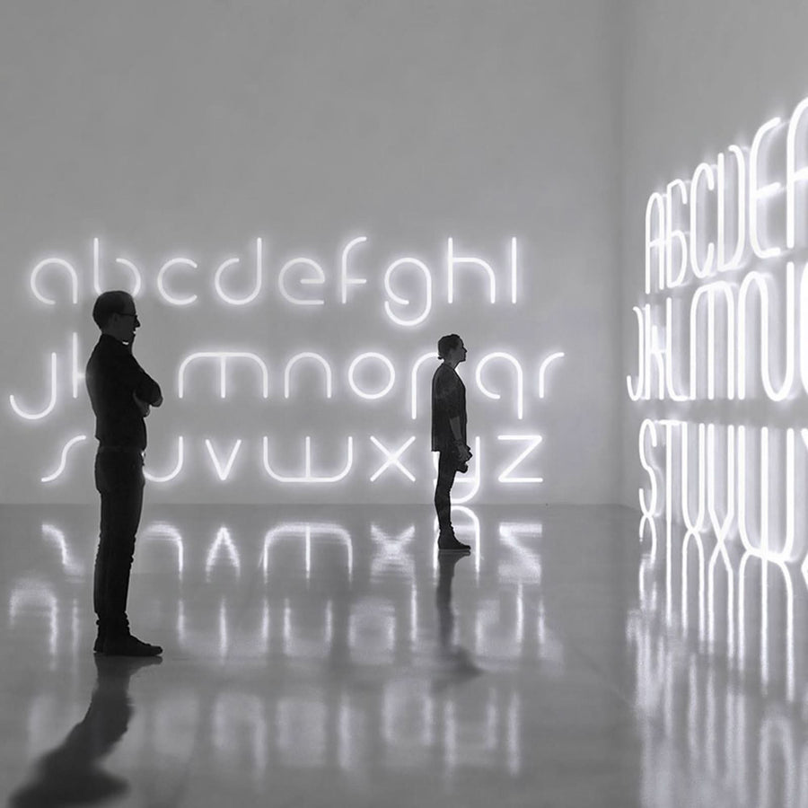 Deux visiteurs contemplent une immense installation de lettres lumineuses blanches avec reflets au sol.
