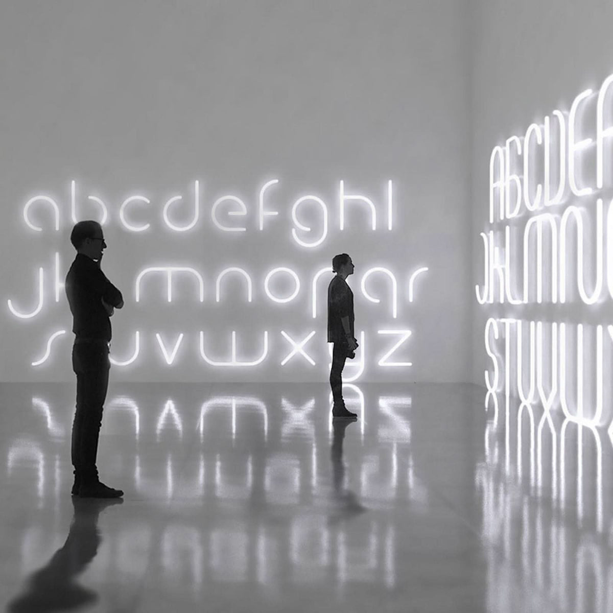 Deux personnes contemplent une grande installation murale de lettres lumineuses.