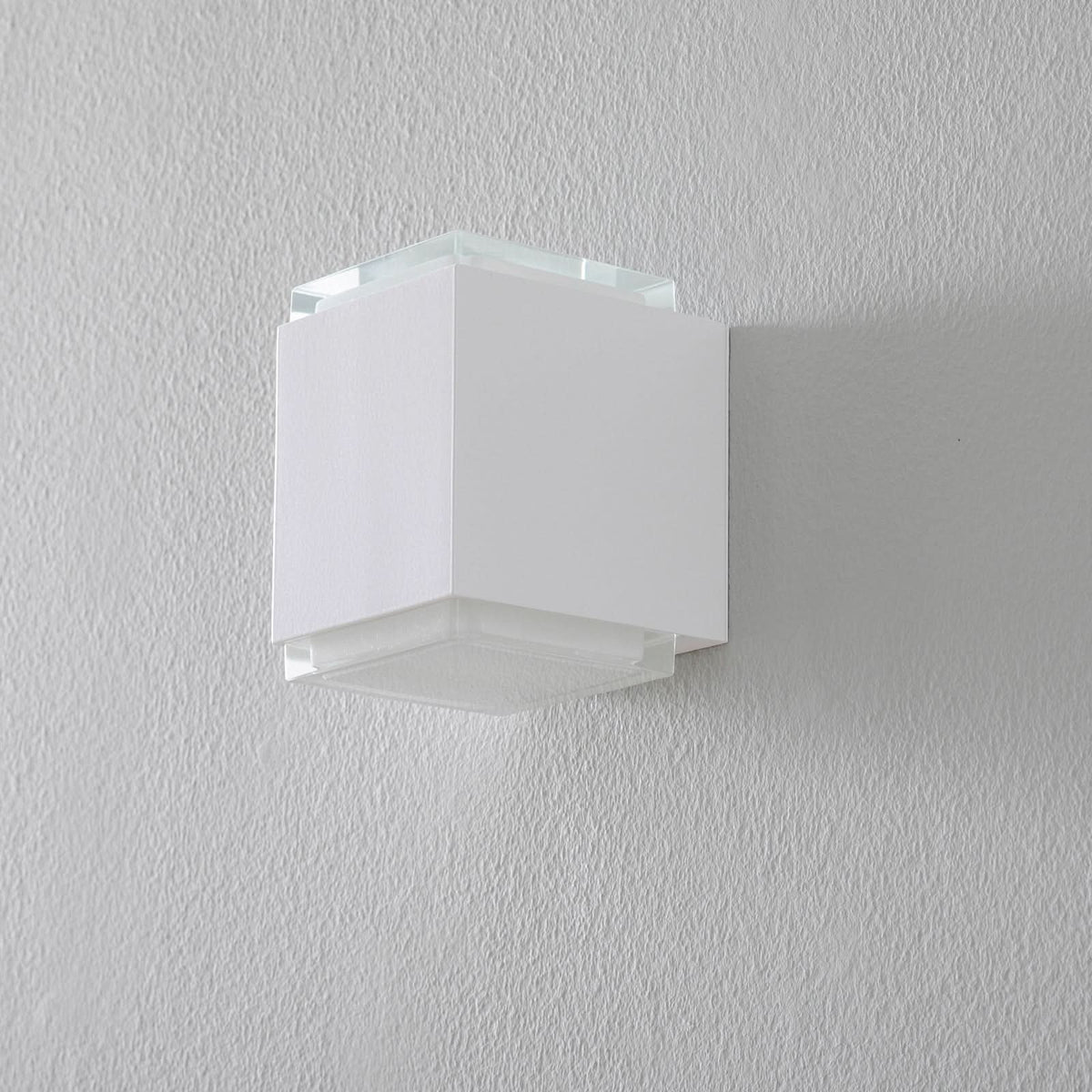 Vue de face du luminaire mural blanc éteint, fixé sur un mur gris clair texturé.