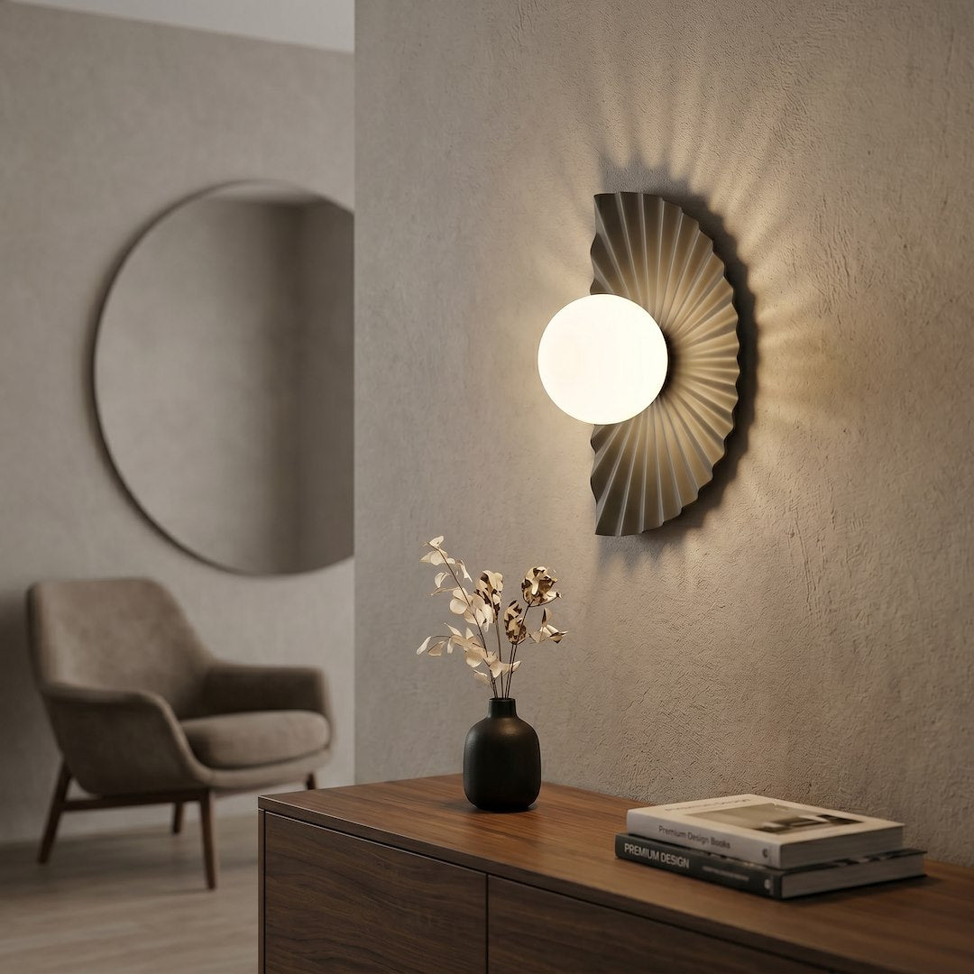 Mise en ambiance : applique allumée, ombres radiales sur mur texturé, vase et livres.