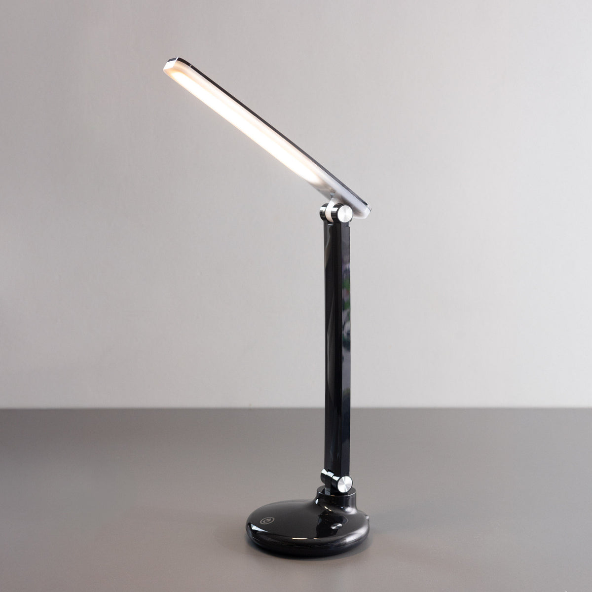 Lampe de bureau noire vue de face, tête inclinée allumée sur fond clair