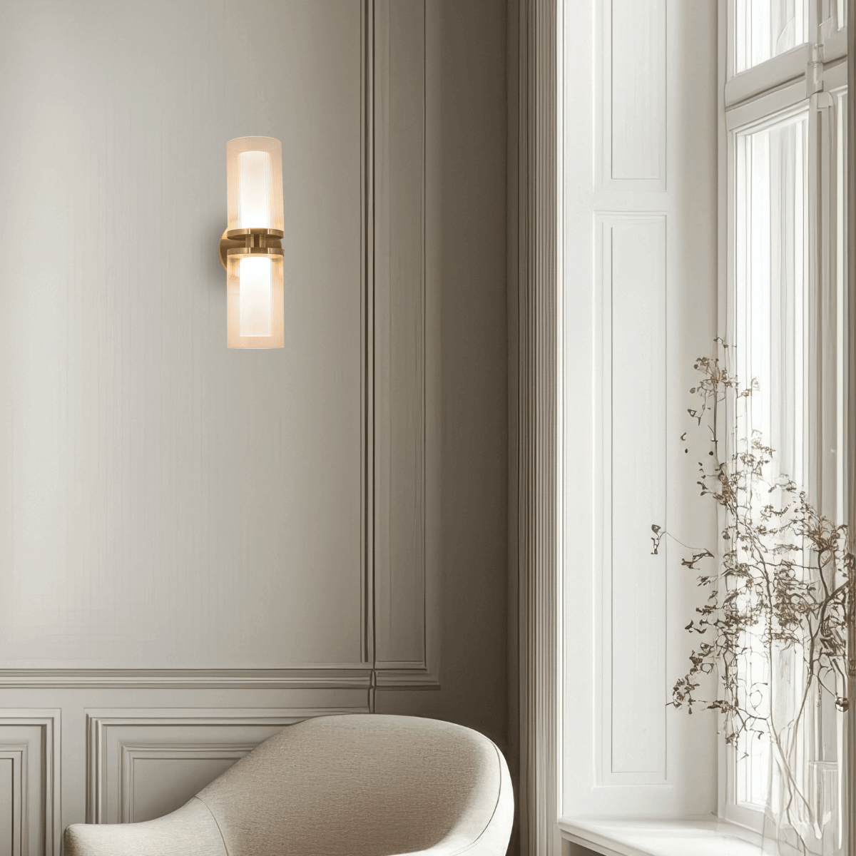 Applique murale dorée allumée sur boiserie grise, avec fauteuil beige et branches séchées près fenêtre.