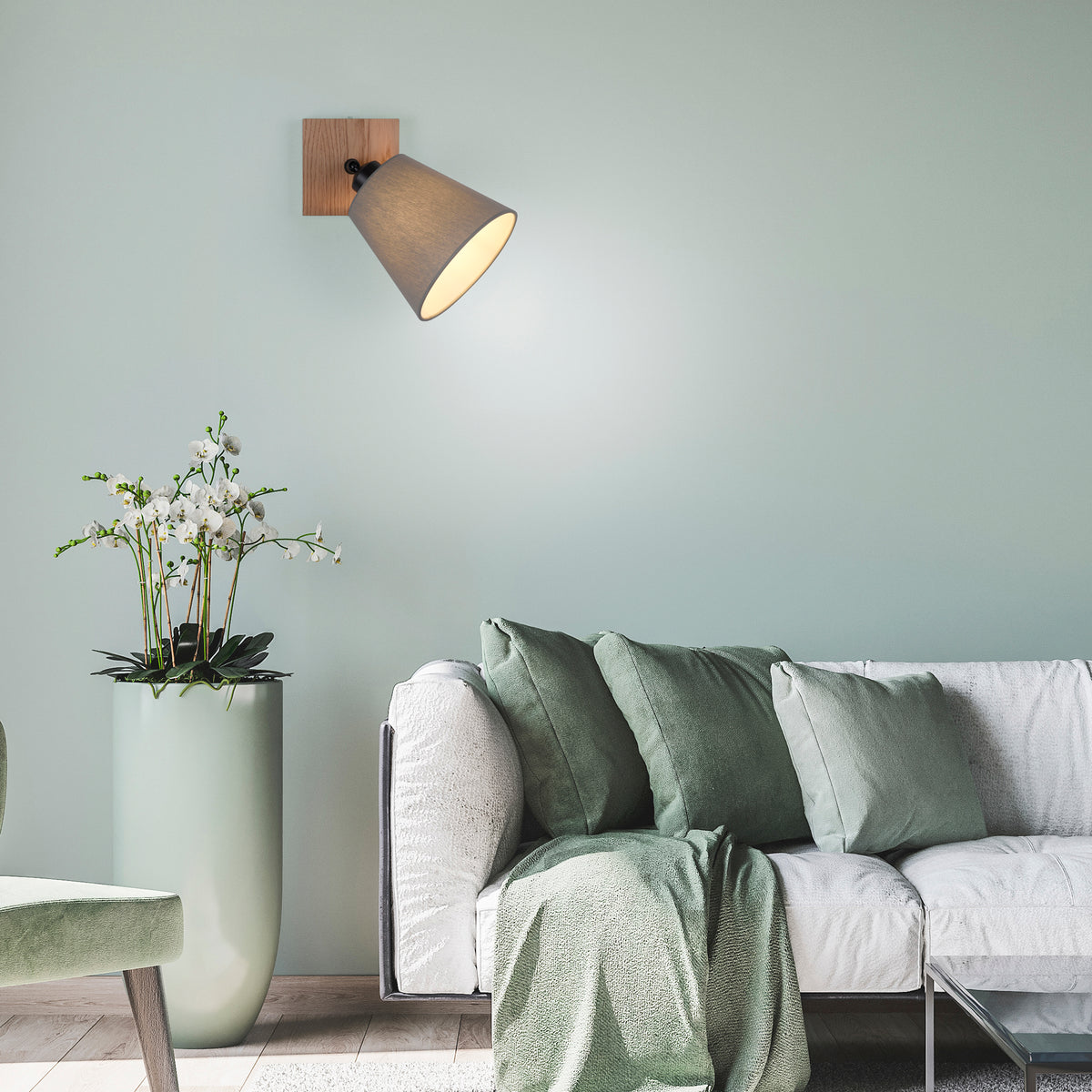 Mise en ambiance : applique murale allumée sur mur vert pâle, canapé et coussins verts, plante.
