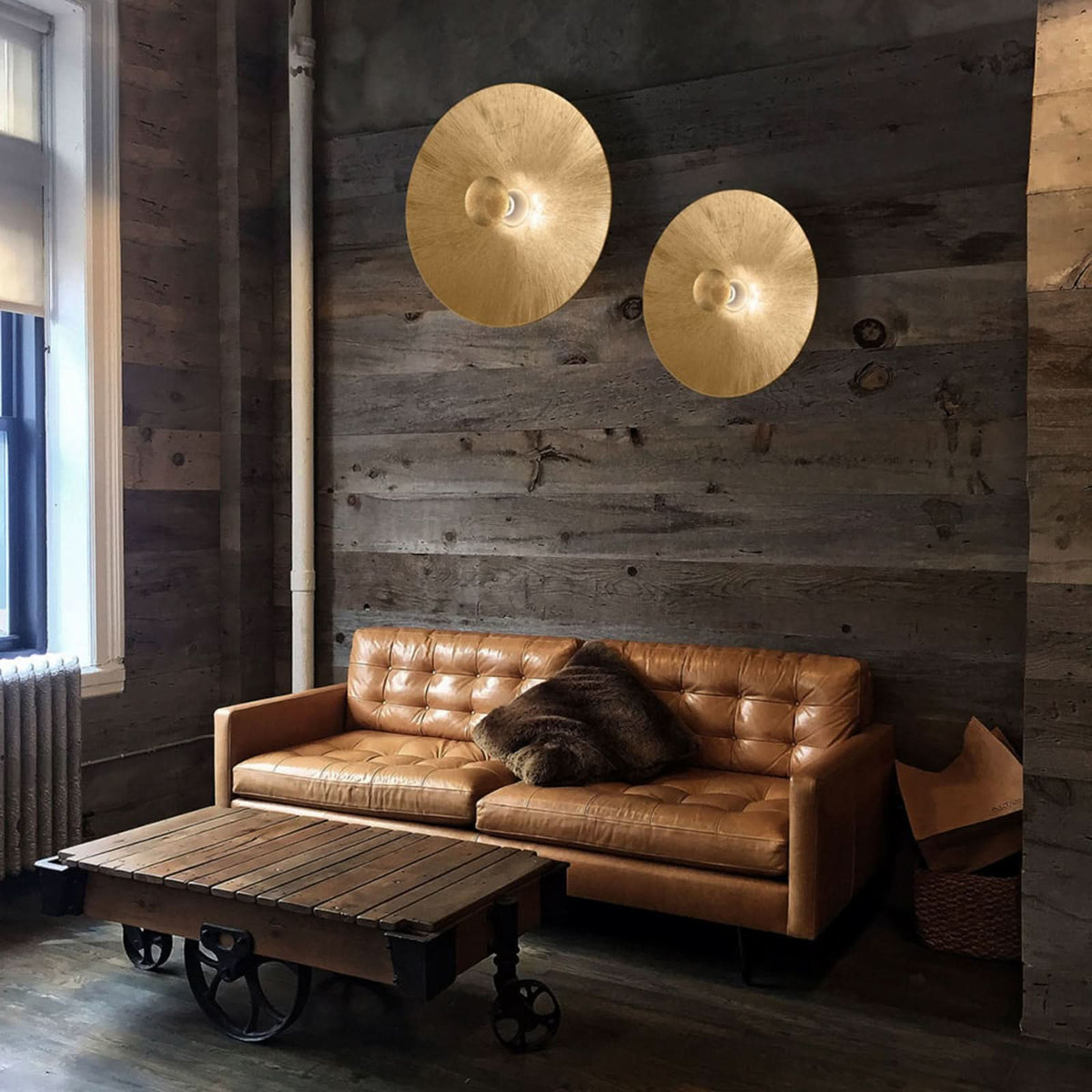 Deux appliques dorées éclairent un mur en bois brut au-dessus d’un canapé en cuir.