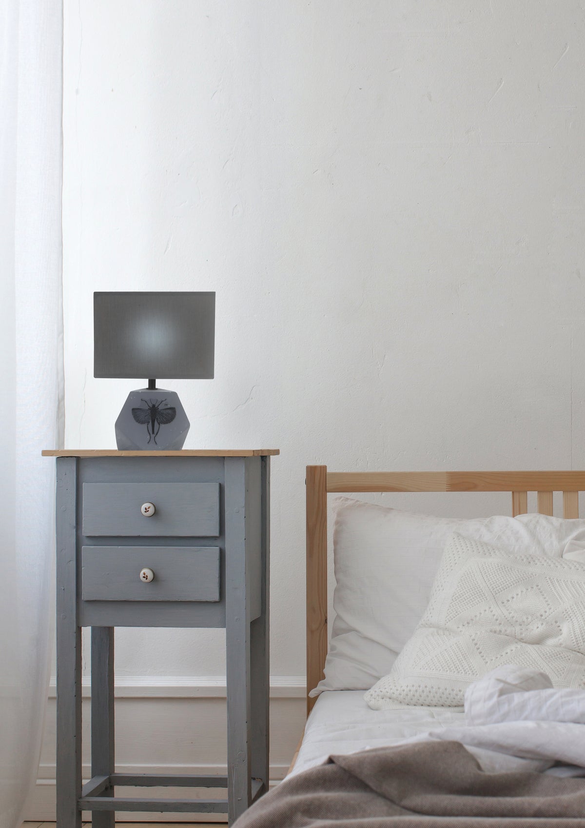 Lampe allumée sur une table de nuit grise, dans une chambre à coucher lumineuse.