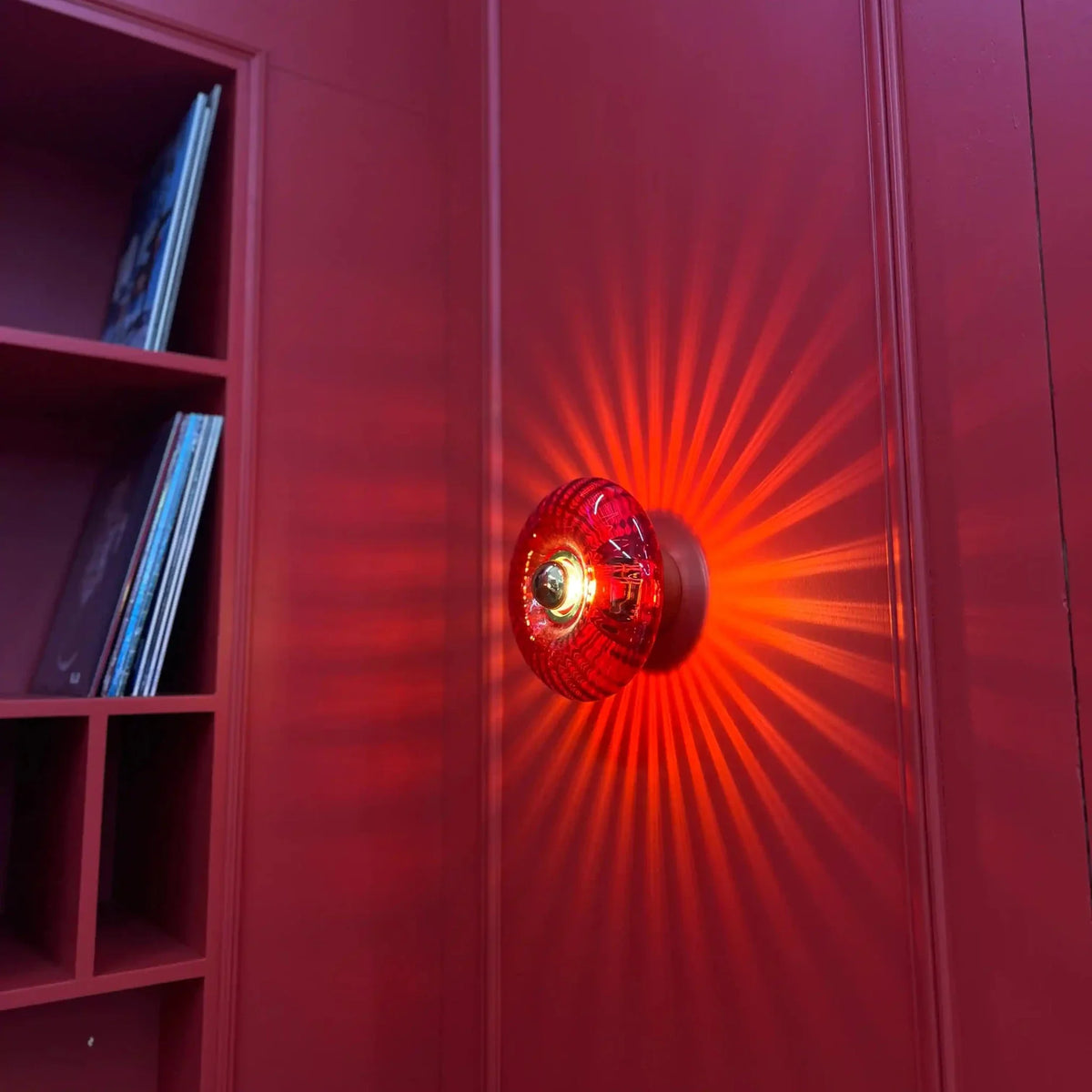 Applique en verre rouge orangé fixée sur un mur bordeaux, lumière incandescente rayonnante