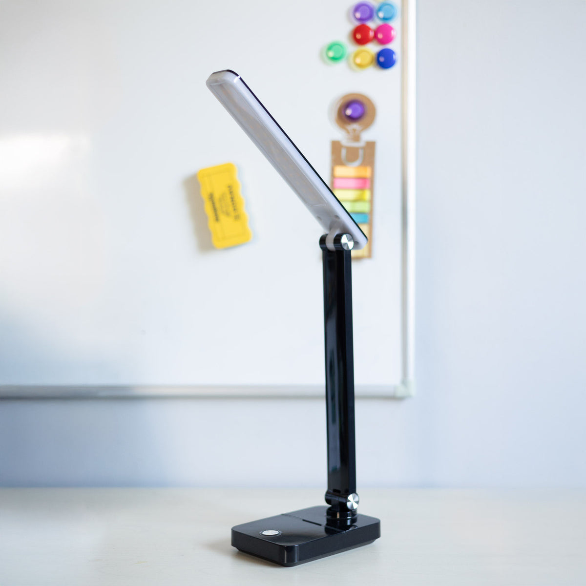 Lampe de bureau noire posée devant un tableau blanc avec fournitures colorées.