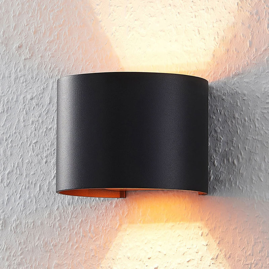 Applique murale noire cylindrique fixée sur un mur texturé blanc, lumière chaude diffusée haut et bas.