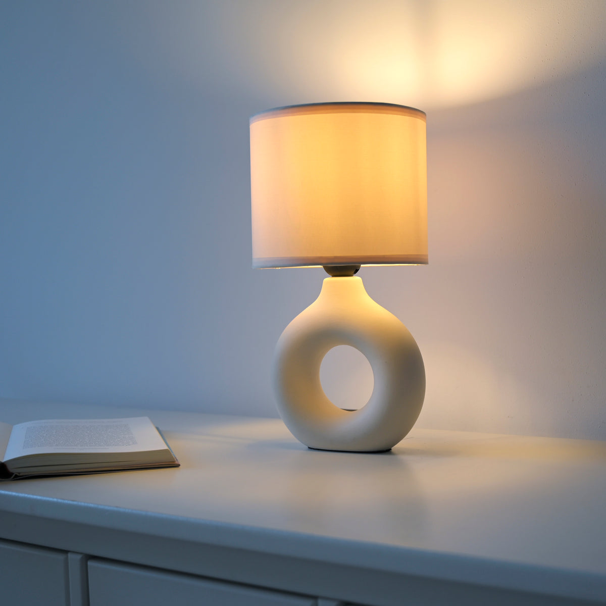 Mise en ambiance nocturne, lampe allumée posée sur meuble avec livre ouvert à côté.