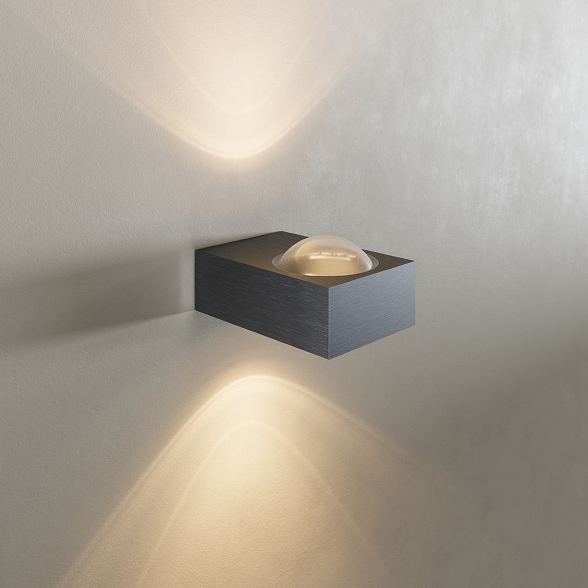 Vue de face sur mur blanc, applique noire brossée diffusant un éclairage haut et bas.