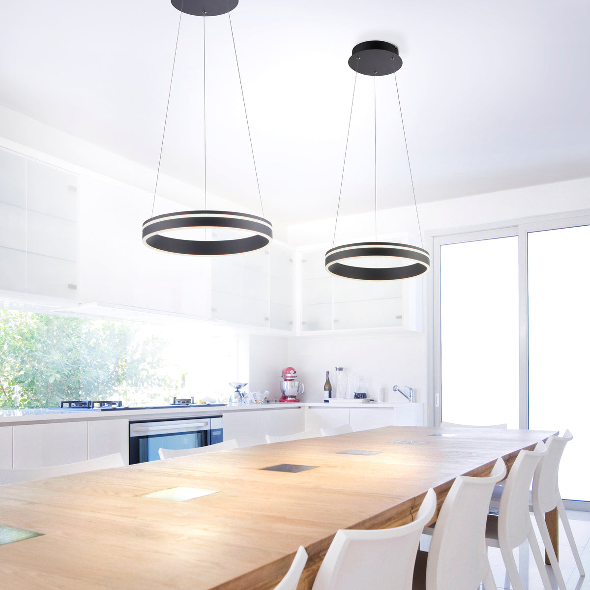 Deux suspensions annulaires anthracite éclairent une cuisine blanche avec une grande table en bois.