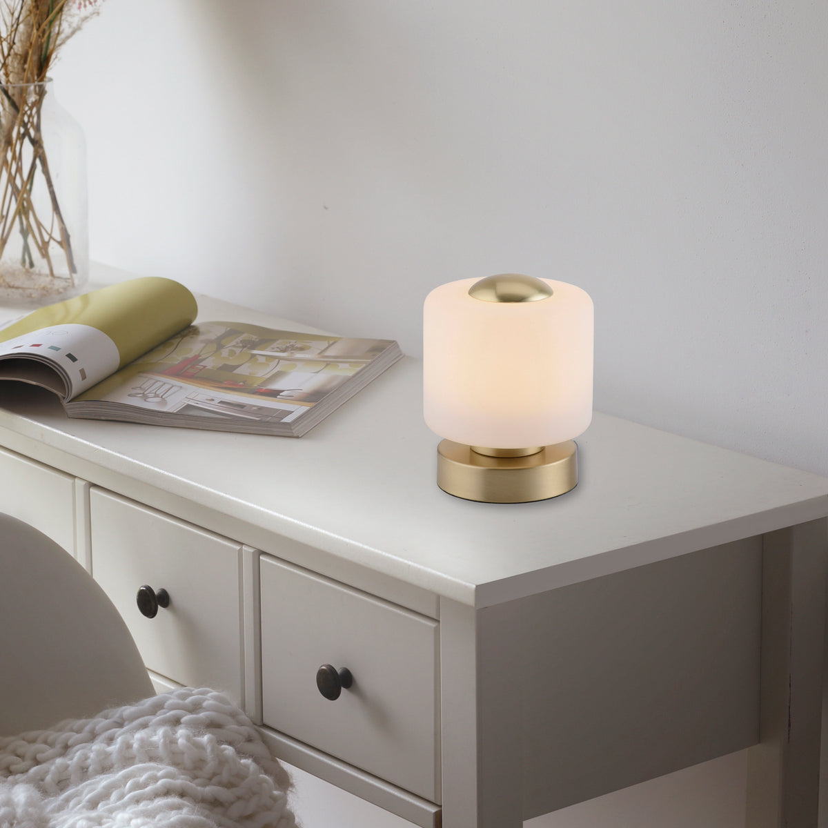 Mise en ambiance sur une commode blanche, lampe allumée près d’un magazine ouvert.
