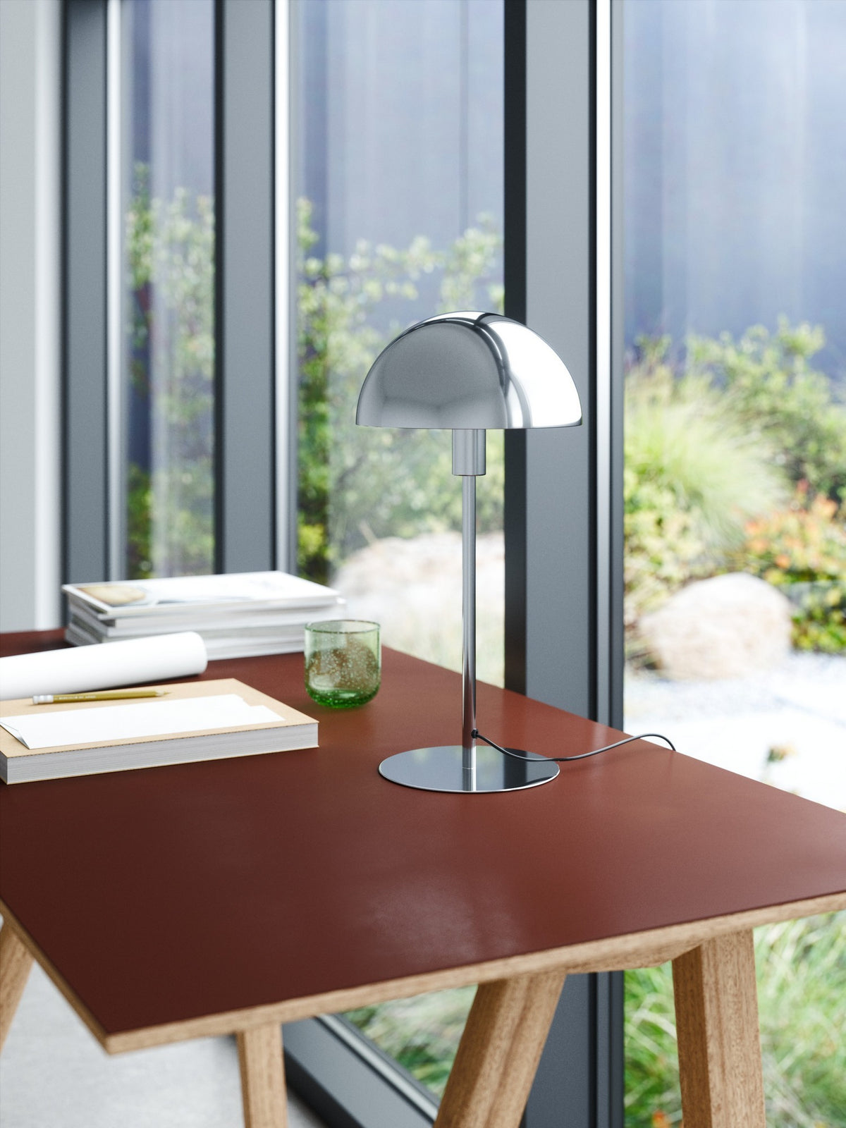 Lampe chromée sur bureau en ambiance, près d'une baie vitrée avec plantes.