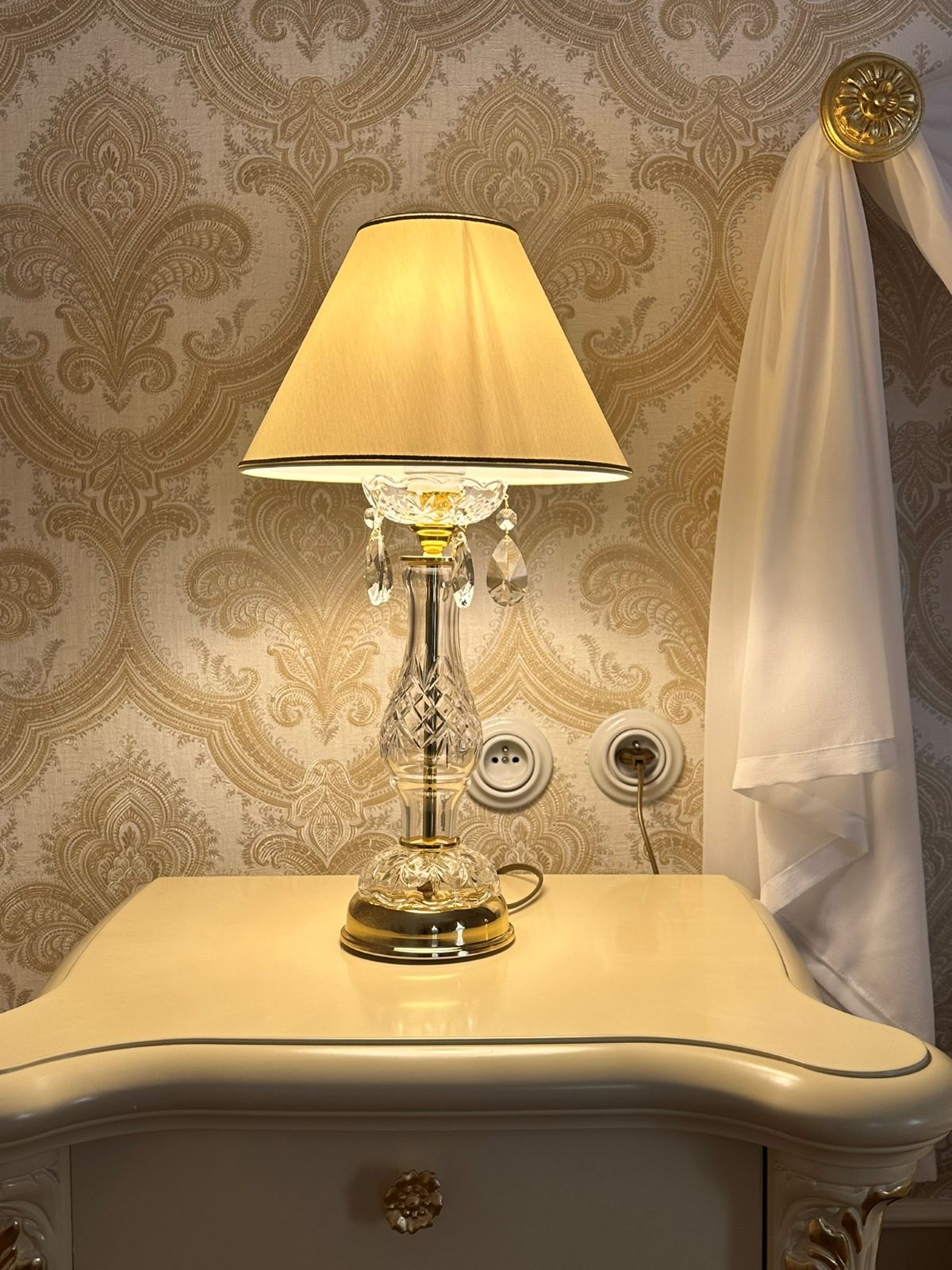 Lampe sur une table de nuit devant papier peint doré, ambiance chambre classique et raffinée.
