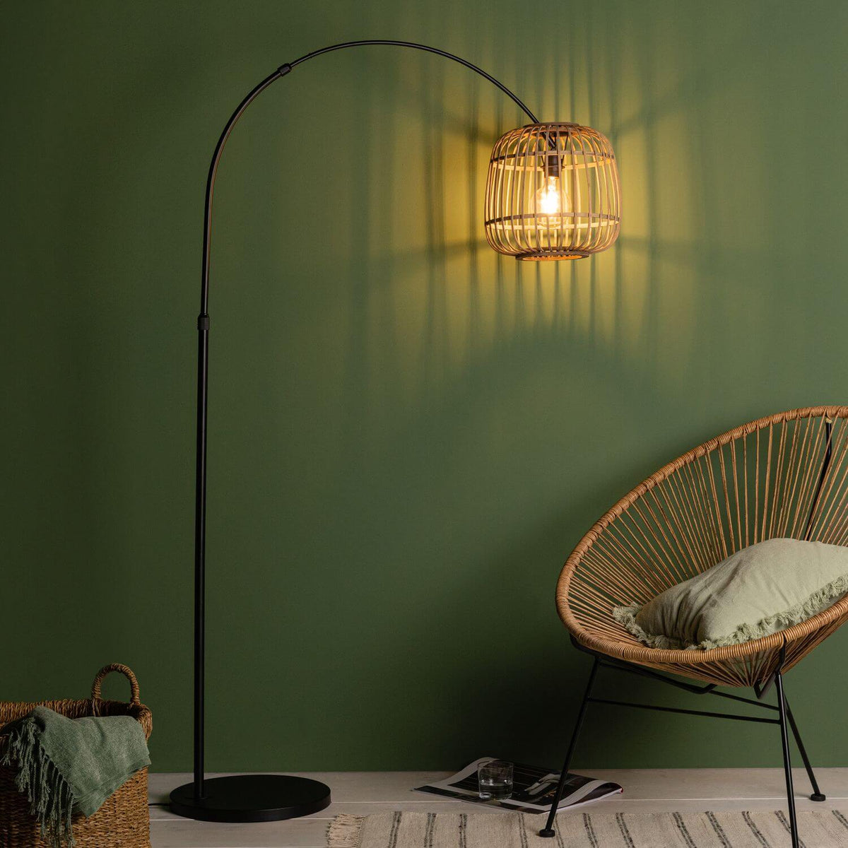 Lampadaire rotin allumé devant un mur vert avec fauteuil en rotin et panier.