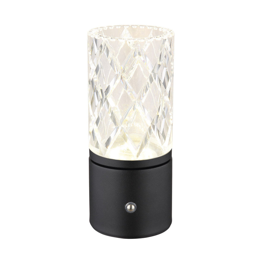 Lampe à poser cylindrique allumée, abat-jour transparent facetté et base noire mate, fond blanc neutre.