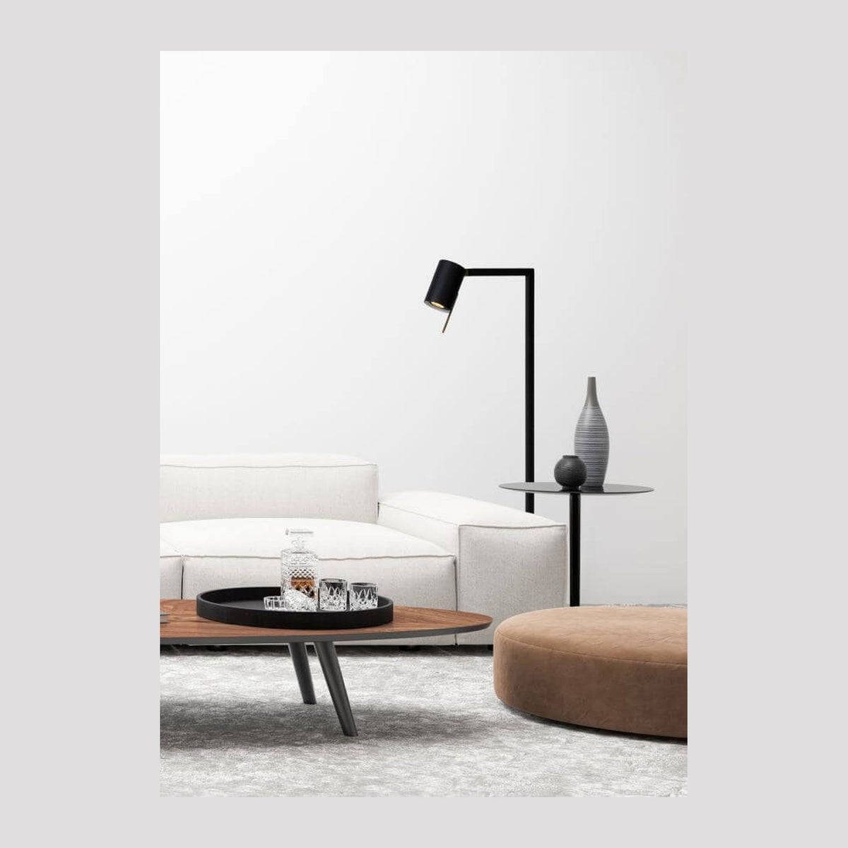 Lampadaire noir installé dans un salon blanc avec canapé, table basse et pouf en ambiance.