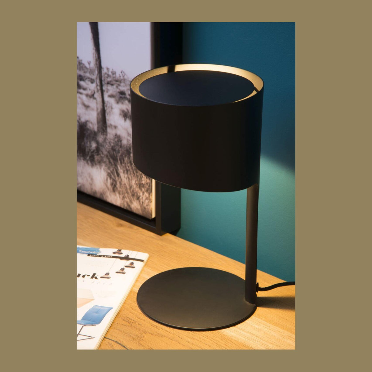 Lampe à poser noire allumée sur un bureau en bois, vue de trois quarts en ambiance.
