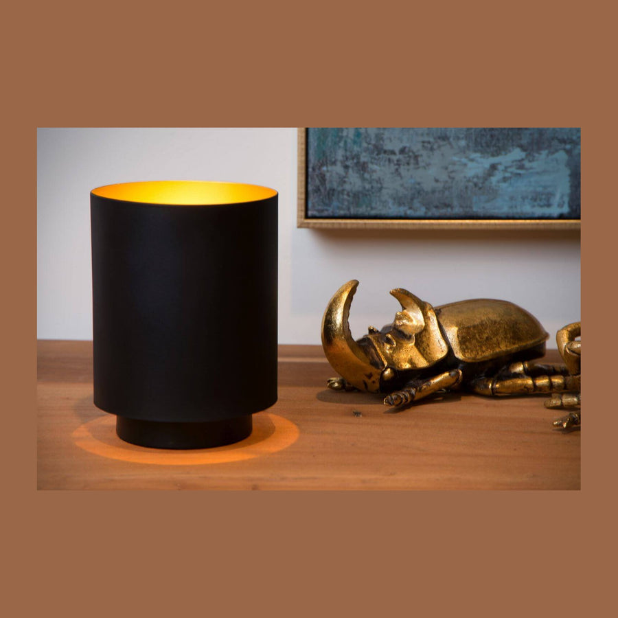 Lampe à poser cylindrique noire et dorée, posée près d'un scarabée doré sur une table.