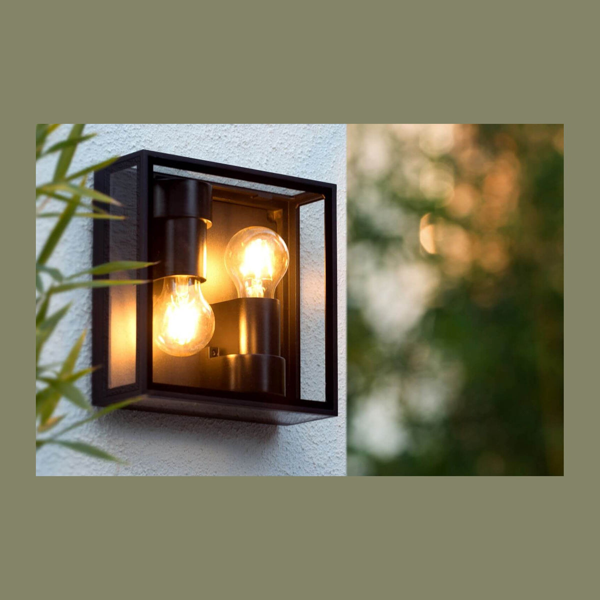 Luminaire extérieur noir fixé au mur, deux ampoules allumées, feuilles de bambou à gauche.