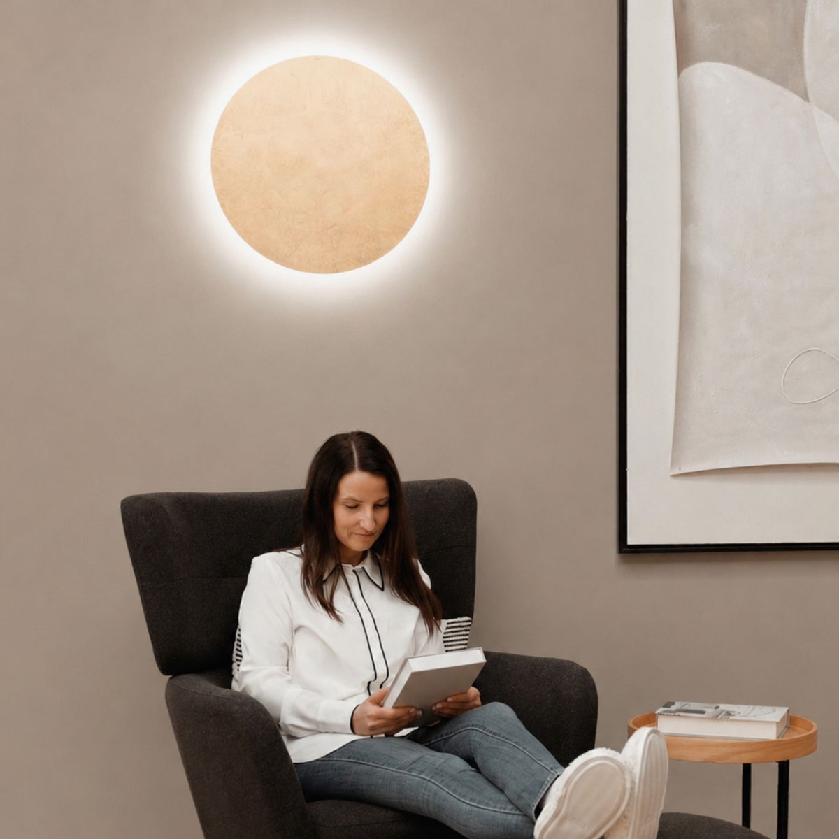Femme lisant dans un fauteuil, une applique ronde dorée crée un halo lumineux sur le mur.