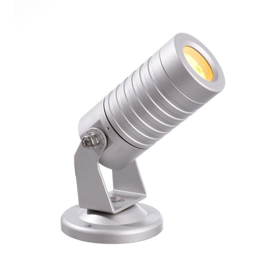 Projecteur extérieur argenté incliné, tête cylindrique nervurée et lentille allumée.