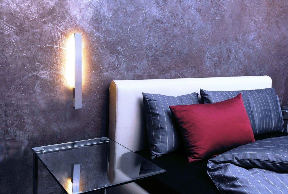 Applique murale allumée dans une chambre, mur texturé, table de chevet et lit en arrière-plan.