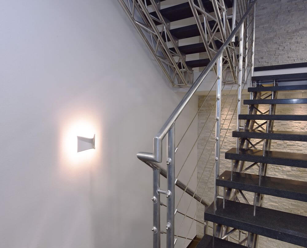 Applique murale allumée sur un mur d'escalier, halo lumineux et rampe métallique.