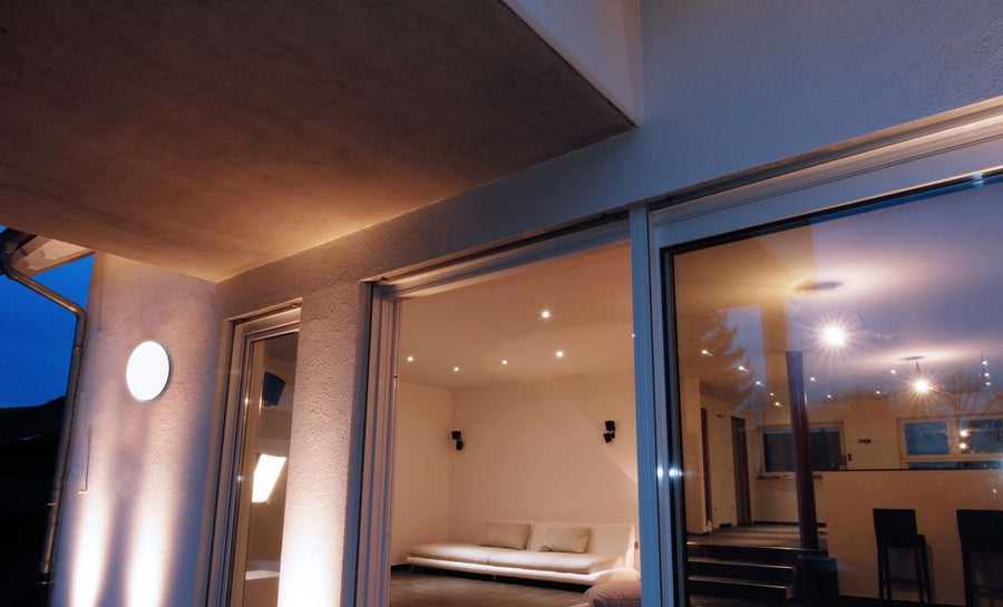 Luminaire extérieur allumé sur une façade, près d’une baie vitrée ouverte sur le salon.
