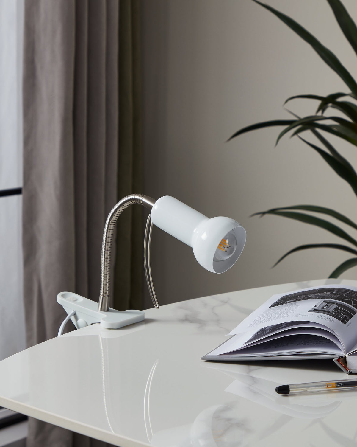 Lampe de bureau blanche allumée, posée sur un bureau blanc, près d'une revue et d'un stylo.