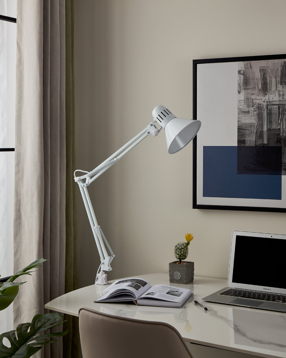 Lampe blanche éclairant un bureau rangé avec livre ouvert, cactus et ordinateur