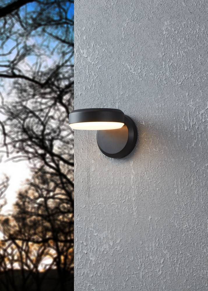 Mise en ambiance extérieure : applique murale noire allumée sur un mur gris texturé et branches floues.