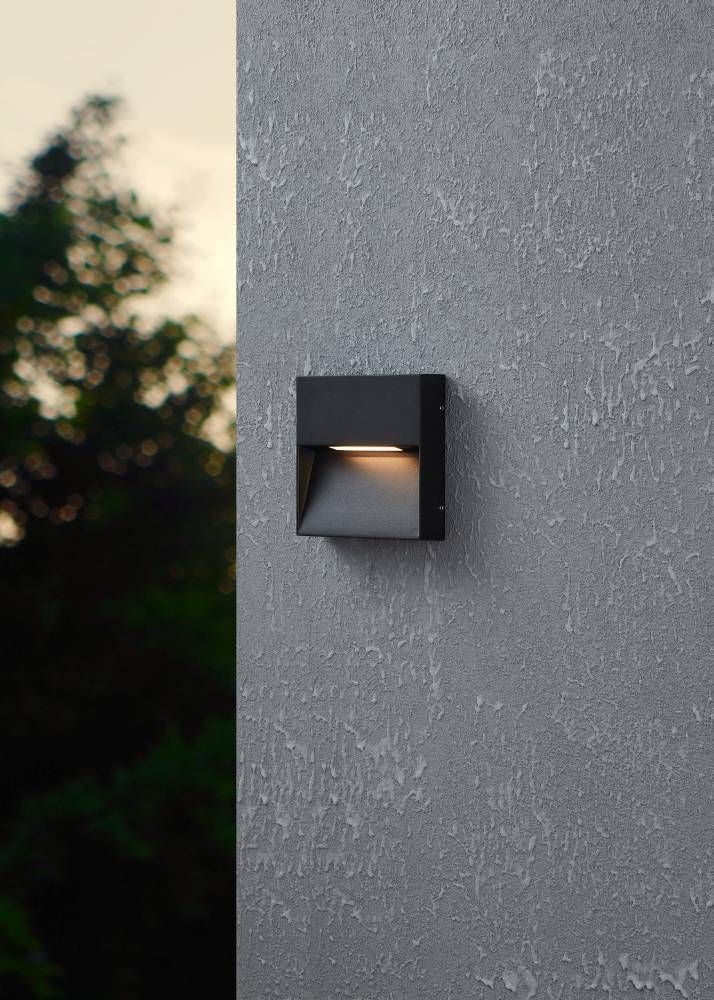 Photo d'ambiance montrant l'applique noire fixée sur une façade grise, lumière allumée