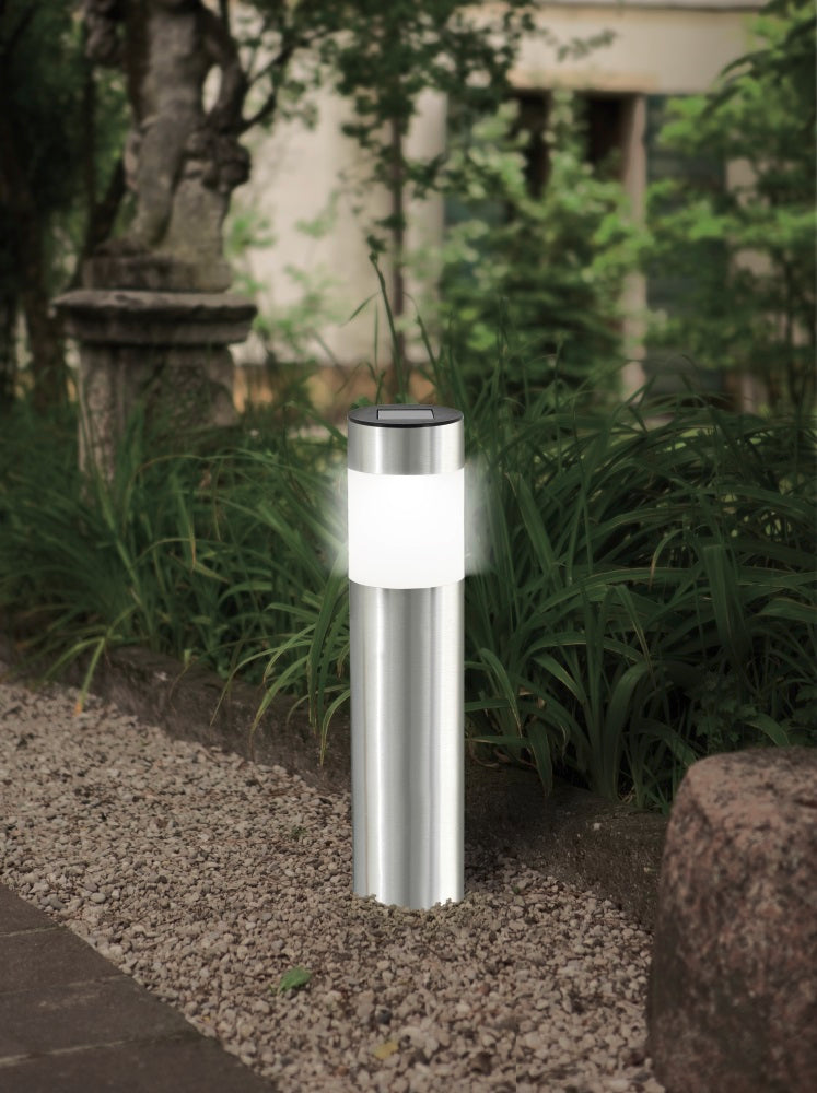 Luminaire inox cylindrique allumé, planté dans gravier près de plantes, en extérieur.