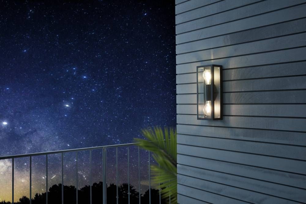 Luminaire mural allumé sur façade claire, ciel étoilé, voie lactée, rambarde et feuillage.