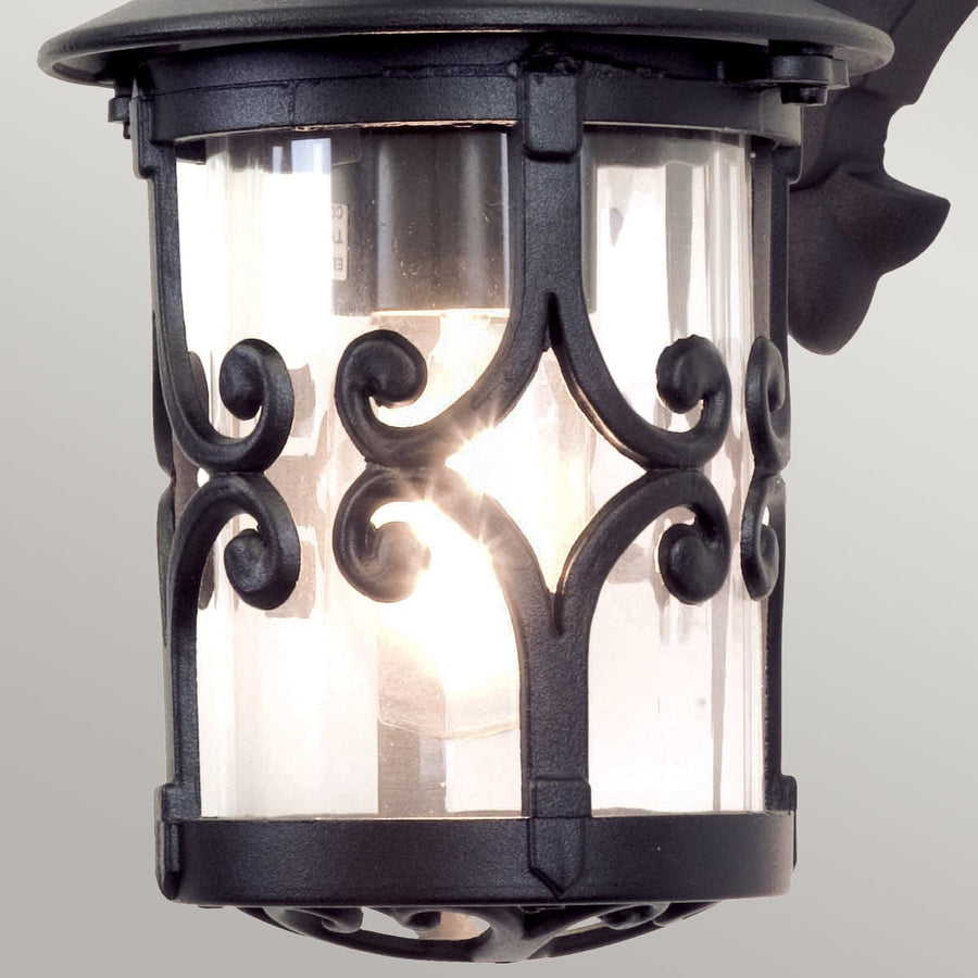 Gros plan de la lanterne : verre transparent, volutes de fer forgé, ampoule allumée