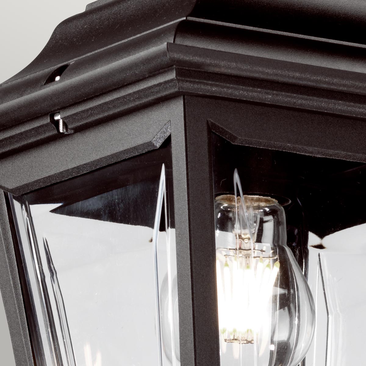 Détail du haut du luminaire : armature noire, verre transparent et ampoule allumée.
