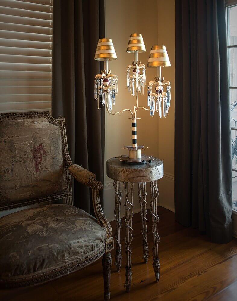 Lampe posée sur guéridon près fauteuil, ambiance intérieure avec rideaux et parquet.