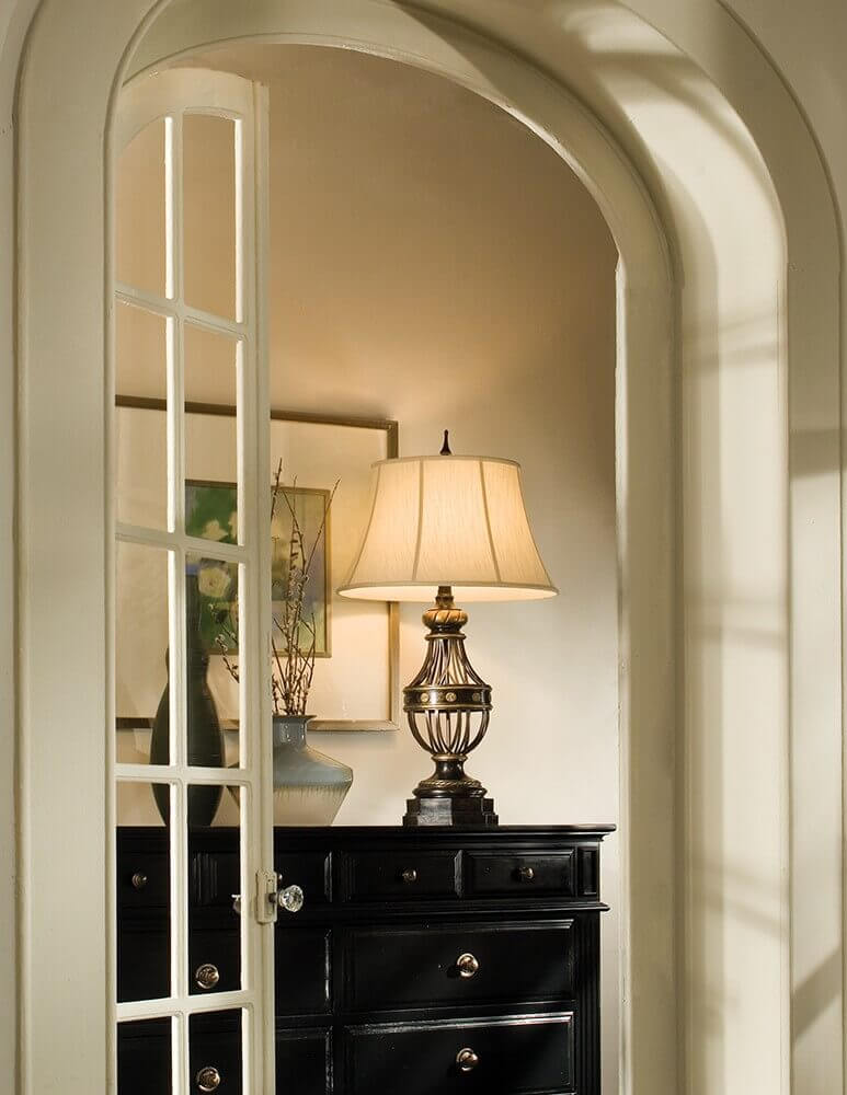 Mise en ambiance de la lampe allumée posée sur une commode noire dans une niche.