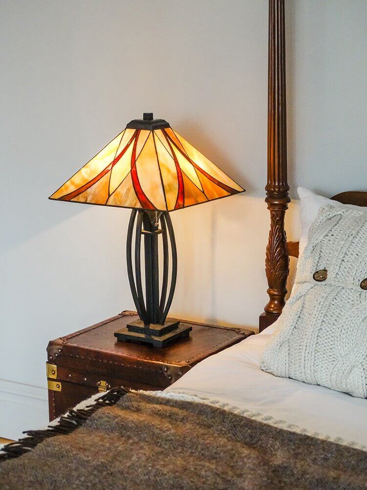 Lampe allumée posée sur une malle près d’un lit, ambiance chaleureuse avec coussin et plaid.