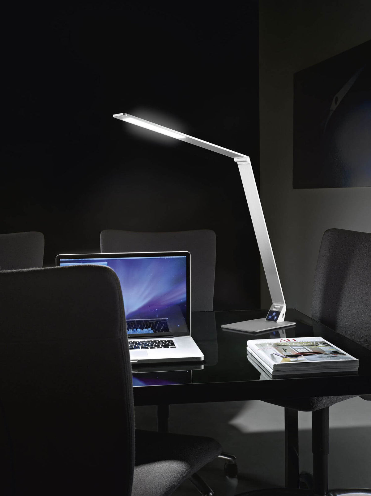 Lampe de bureau allumée sur table noire dans un bureau sombre, ordinateur portable et revues.