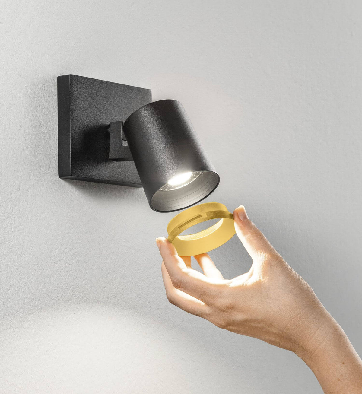 Main présentant la bague dorée devant le spot mural noir allumé