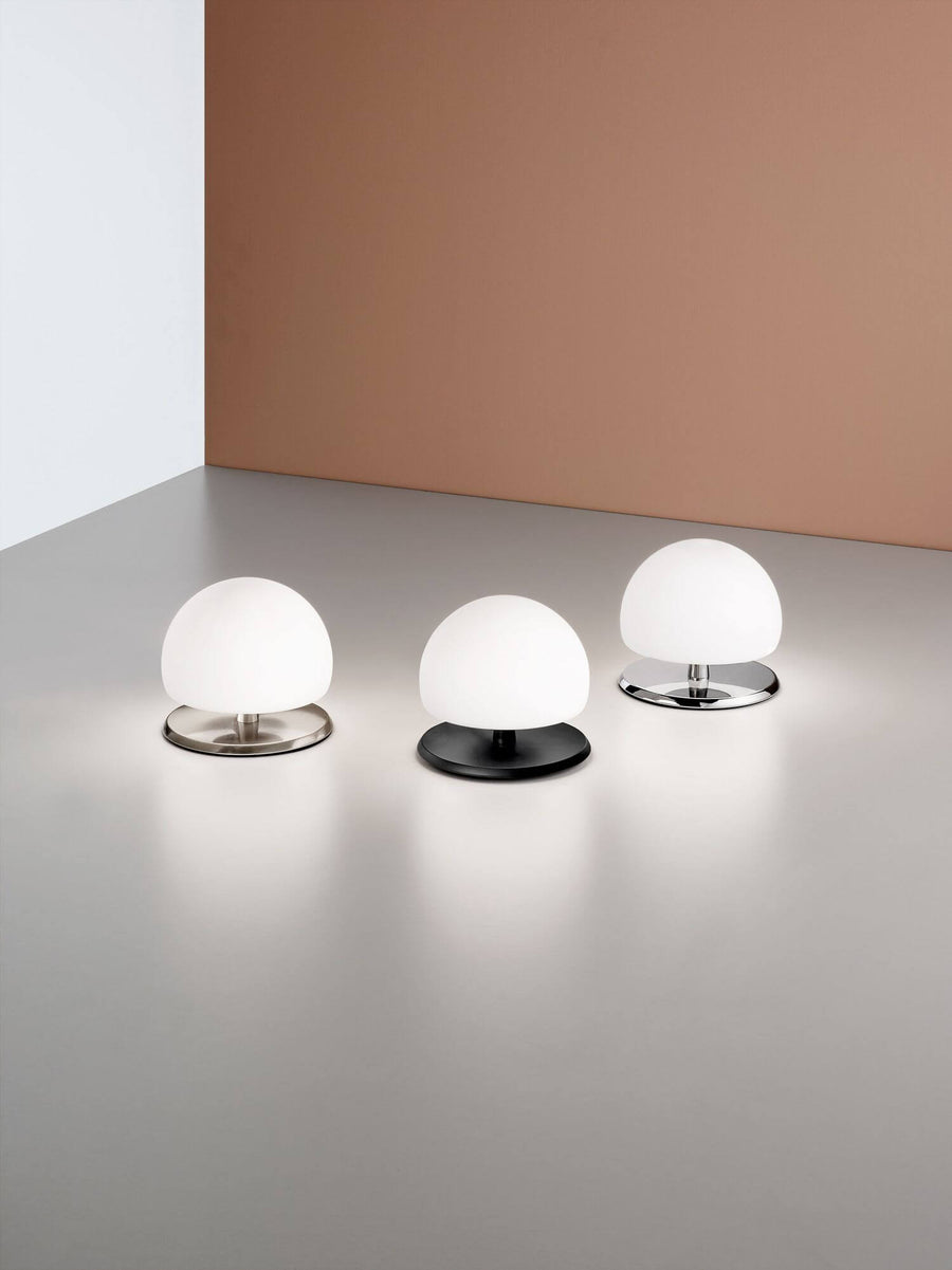 Trois exemplaires de la lampe alignés sur un plan gris, bases chromée, noire et nickel.
