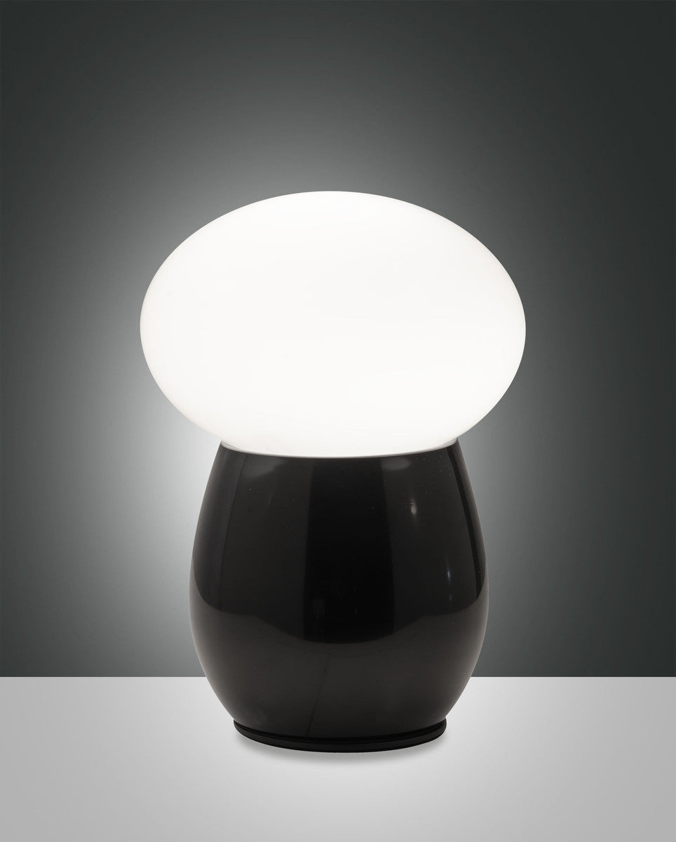 Lampe à poser allumée, vue de face, base noire brillante et diffuseur ovale blanc