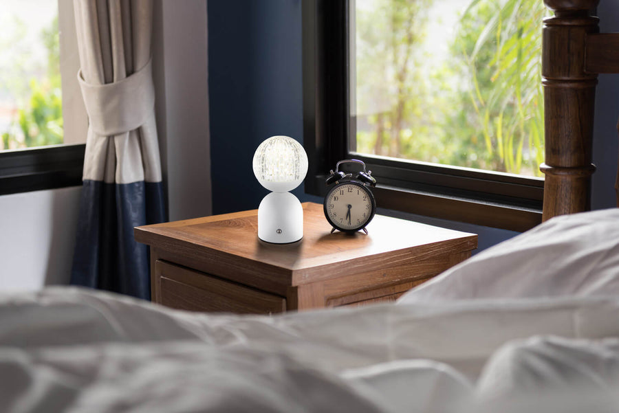 Lampe à poser blanche allumée sur une table de nuit en bois près du lit.
