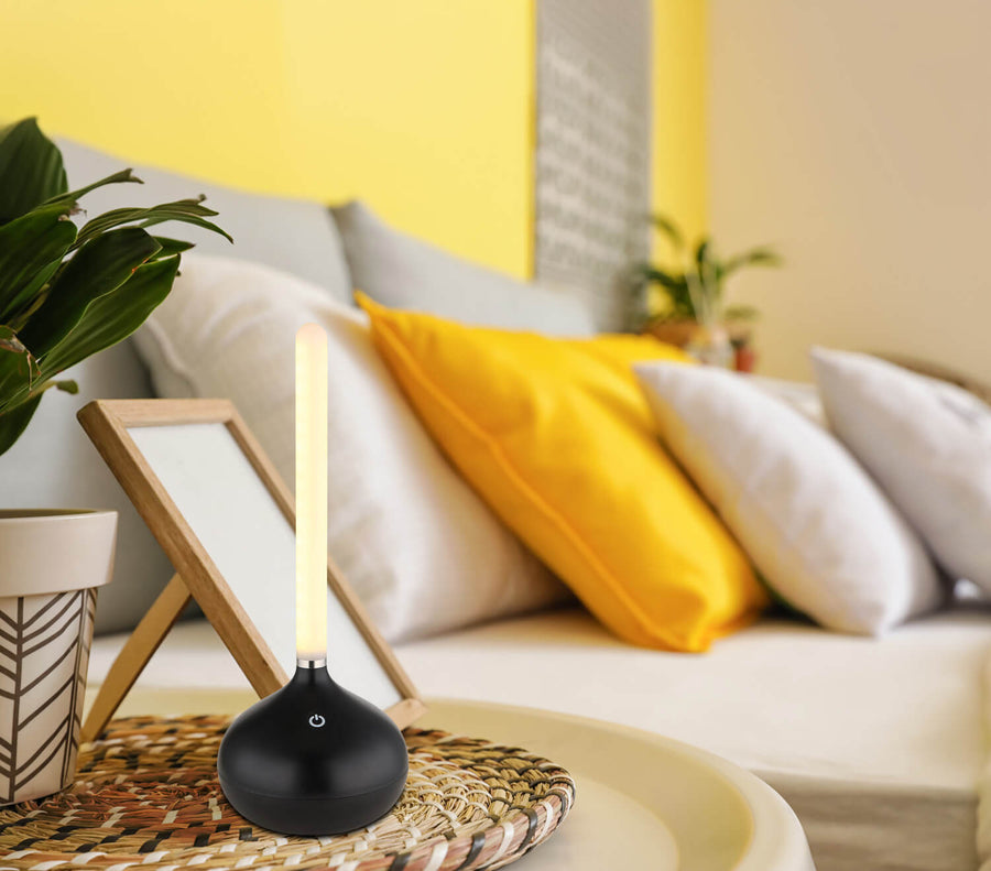 Lampe à poser noire allumée sur une table basse en rotin, canapé jaune en arrière-plan.