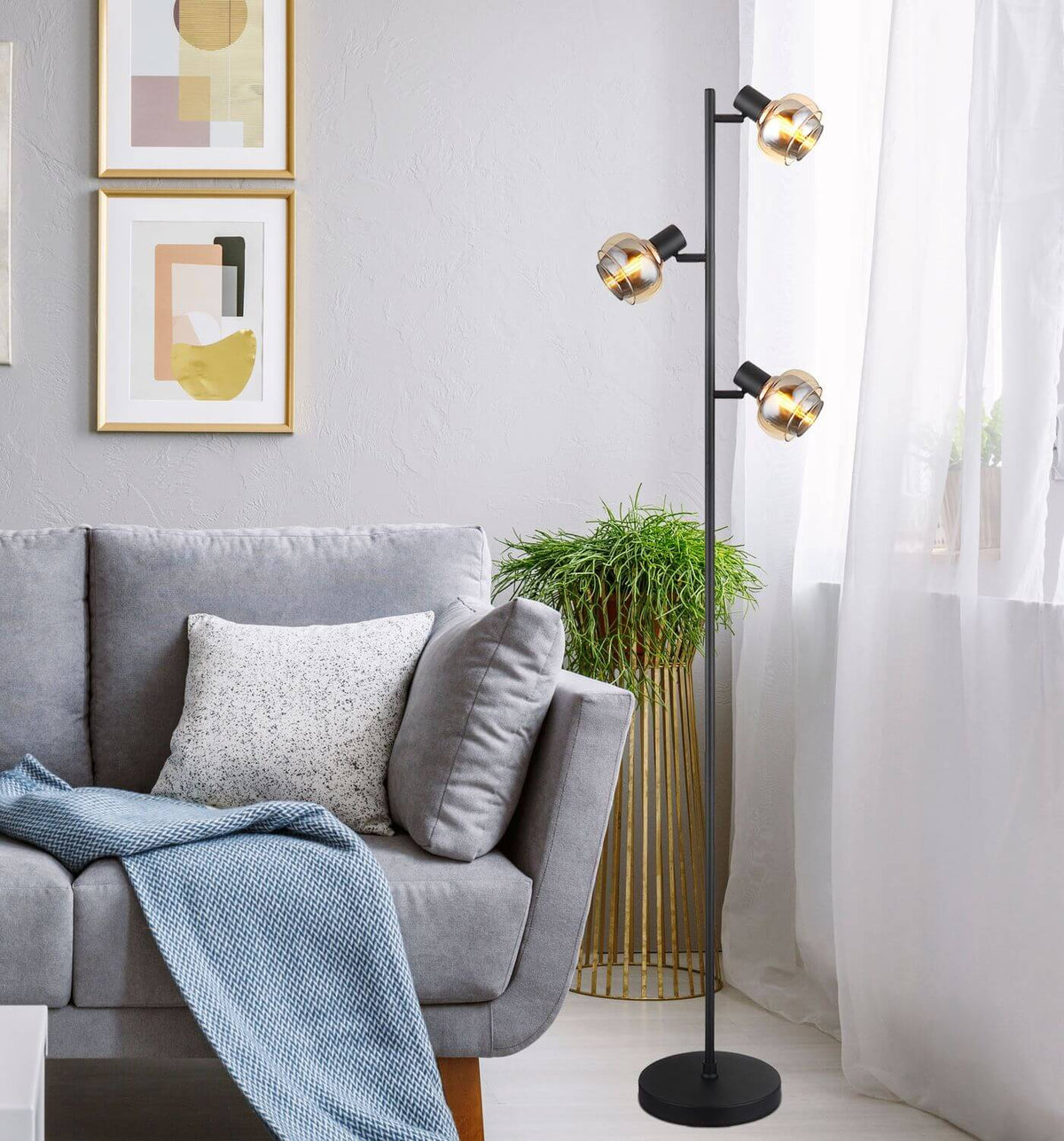 Mise en ambiance du lampadaire allumé près d'un canapé gris, avec rideaux, plante et cadres.